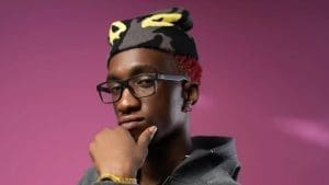 A person wearing glasses and a hat with yellow eyes poses against a pink background, resting their chin on their hand—reflecting the creative spirit of WETALKSOUND and Africa’s Sonic Future Artists.