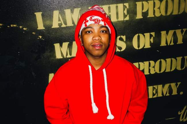 A person in a red hoodie and red beanie stands in front of a black wall with yellow stenciled text, channeling the bold style of independent artist Mfana Kah Gogo from the Amapiano scene.