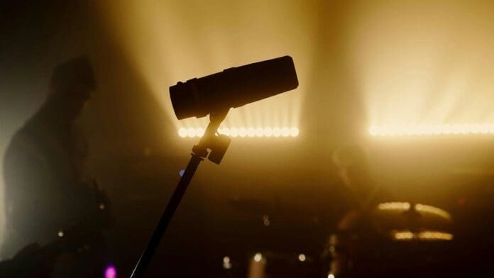 A microphone on a stand is silhouetted against bright stage lights, with blurred musicians and a drum set in the background—setting the scene for protest music or revolutionary rap performances reminiscent of artists like Falz.