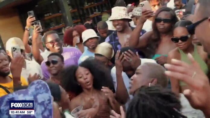 A large group of people dance and celebrate outdoors during the day in DC, some filming with their phones. The FOX LOCAL news logo and current time and temperature appear in the corner.