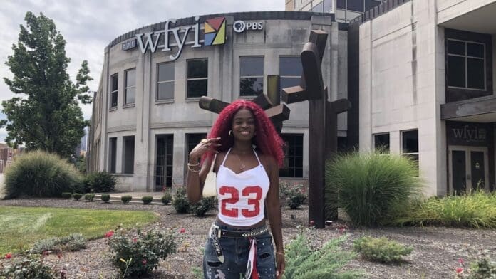 A Haitian American vocalist with red hair stands outside the WFYI PBS building, wearing a white tank top with the number 23 and ripped jeans, surrounded by landscaping and greenery.