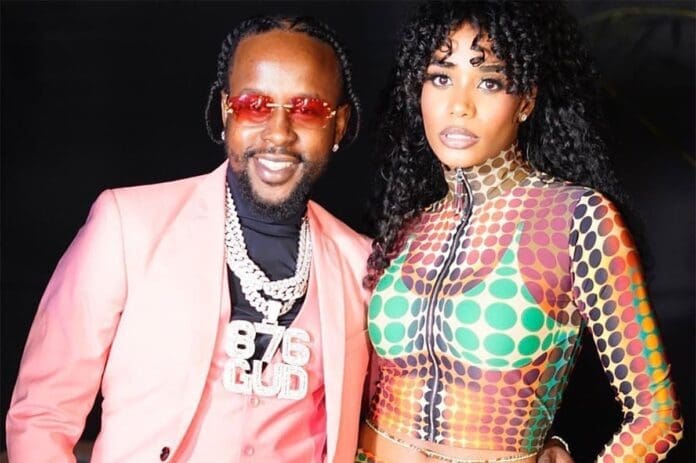 A man in a pink suit with layered chains and Toni-Ann Singh in a colorful patterned outfit pose together against a dark background.