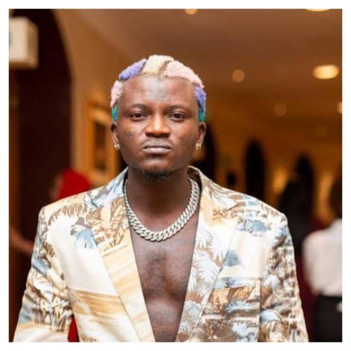A man with multicolored hair, a silver chain necklace, and a patterned blazer stands indoors, looking directly at the camera—a style reminiscent of Burna Boy.