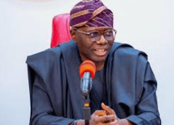 A man wearing glasses and traditional attire speaks into a microphone at a table, sitting against a plain background during an event with Lagos govt officials ahead of AFRIMA.