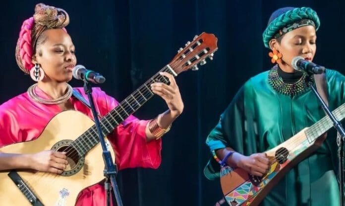 Two women in colorful outfits and headwraps play guitars and sing into microphones during the vibrant opening performance at the Sankofa Heritage Festival, celebrating the rich rhythms of Music In Africa.