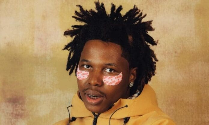 A man with short curly hair wears a yellow jacket and has pink patterned adhesive strips under his eyes, posing in front of a textured beige background—perfect for a Spotify feature on Africa’s rising artists or the NXT playlist.