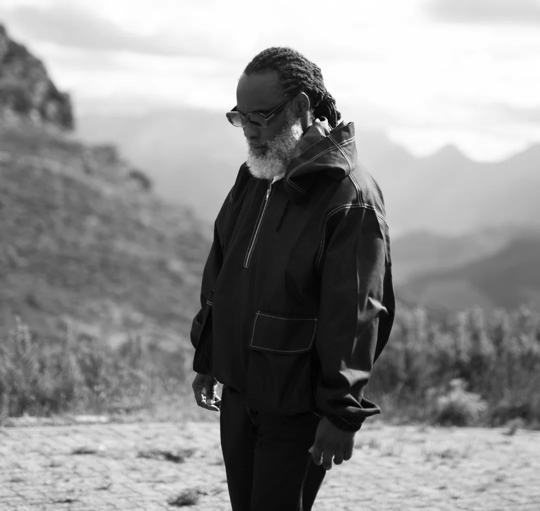 A man with glasses and a beard stands outdoors in a mountainous landscape, wearing a dark jacket, channeling the soulful energy of Nigerian artist Afẹ́fẹ́ Ìfẹ's new single.