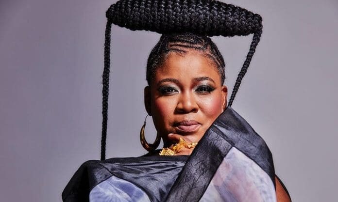 A woman with braided hair styled in a sculptural updo, wearing a patterned wrap and gold jewelry, poses against a neutral background—radiating the elegance often seen at the 2026 South African Music Awards.