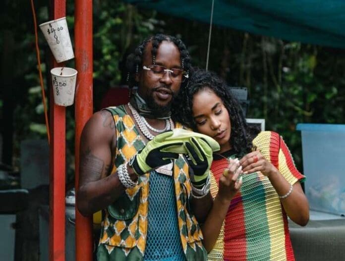 Two people stand outdoors; one, channeling Popcaan's laid-back vibe, holds a green plantain and wears gloves, while the other looks closely at something in her hands. Cups with prices hang on a pole beside them.