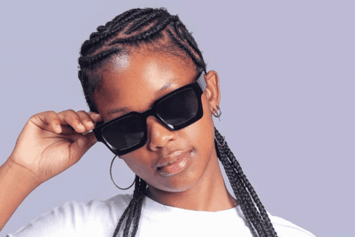 A person with braided hair adjusts large black sunglasses with one hand, wearing a white shirt and hoop earrings against a light purple background, embodying the vibrant style often seen in Afropop culture from Eswatini.