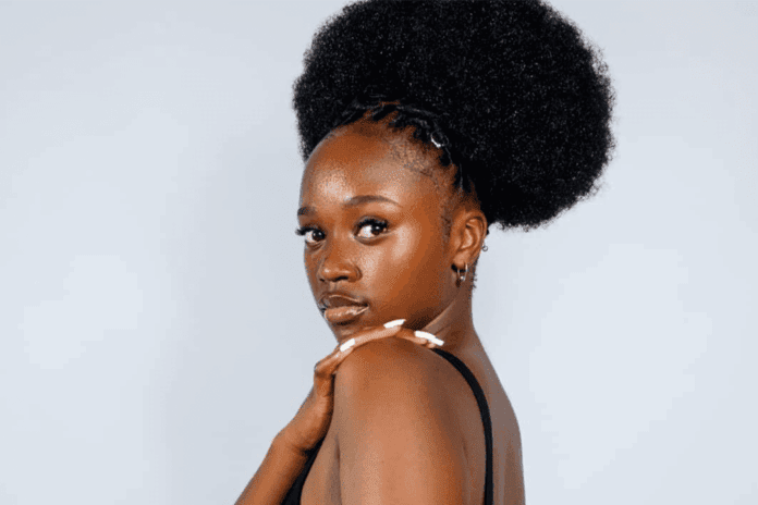A woman with dark skin and a large, round afro poses against a plain light background, looking over her shoulder with a neutral expression, embodying the soulful spirit of Amapiano music.