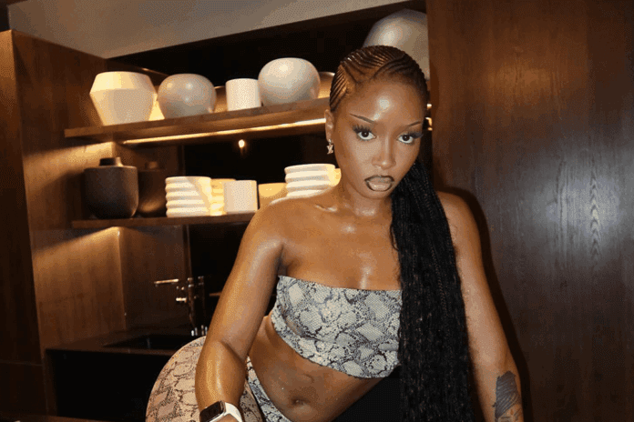 UNCLE WAFFLES, a woman from Eswatini with long braided hair, poses in a modern kitchen wearing a snakeskin-patterned strapless outfit. Wooden cabinets and shelves displaying dishes add to the stylish setting.