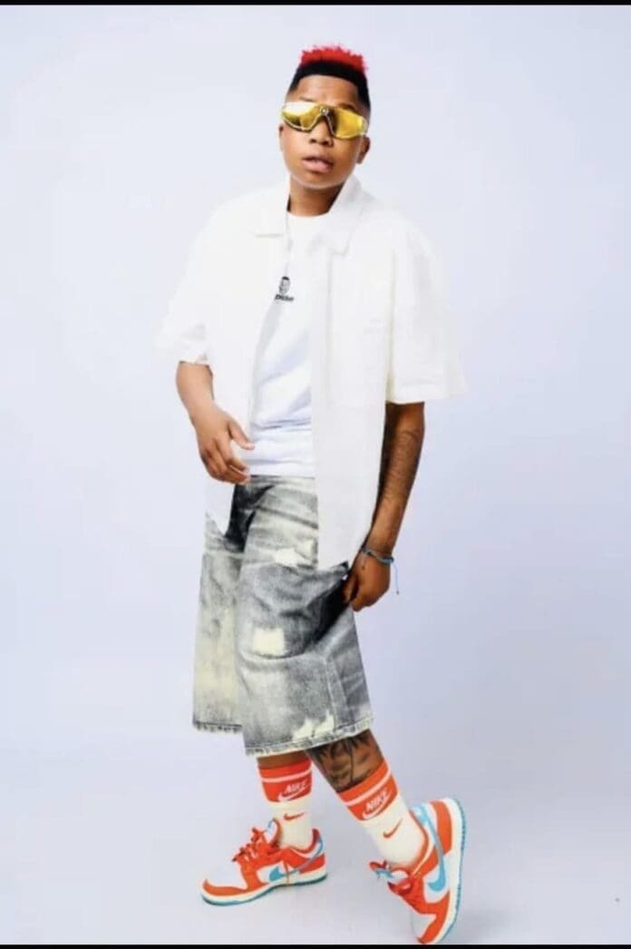 Person wearing a white shirt, distressed denim shorts, orange and white Nike sneakers, tall matching socks, gold sunglasses, and sporting short red hair poses against a plain background with cool Amapiano vibes.