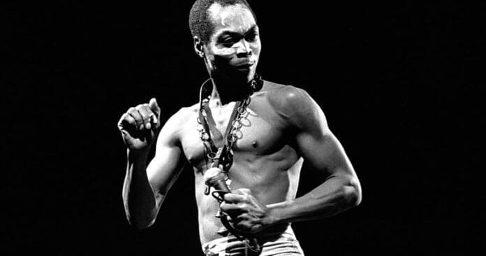 A shirtless man with face paint and a necklace stands against a black background, holding a microphone and looking to the side, channeling the spirit of Afrobeat pioneer Fela Kuti.