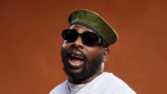 A bearded man wearing sunglasses, a green beret, a white shirt, and a necklace poses with his mouth open against a plain brown background, channeling the vibrant energy of Afro music.