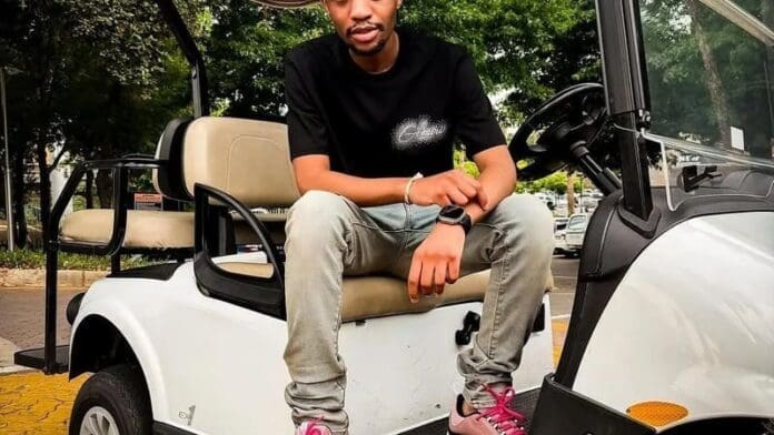 A man wearing a black t-shirt, gray jeans, and pink sneakers sits on the front of a white golf cart parked outdoors, vibing to amapiano hits like "Shela.