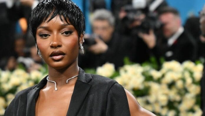 A woman in a black outfit with short black hair poses on a red carpet, channeling Afrobeats glamour; photographers and yellow flowers blur in the background.