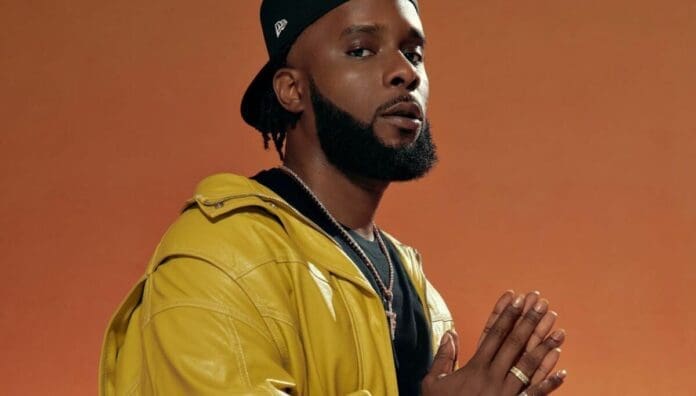 A man with a trimmed beard wearing a yellow jacket and black cap poses with hands together against an orange background, channeling 2000s Afropop vibes reminiscent of Maleek Berry's debut album style.