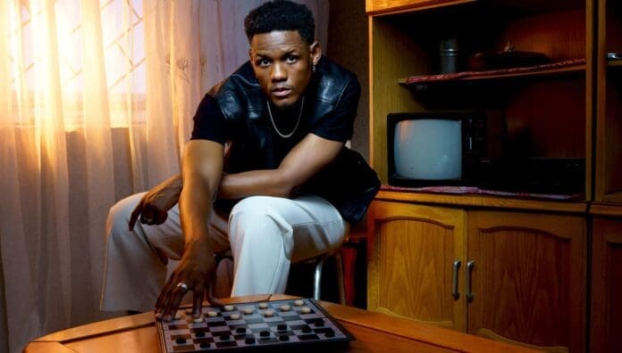 A man in a black shirt and white pants sits beside a checkerboard, reaching for a piece, in a room with wooden furniture and an old television—evoking the soulful vibe of South African artist Samthing Soweto featured on OkayAfrica.