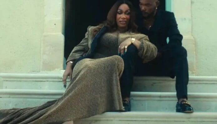 A woman in a long gold dress and a man in a black suit sit together on stone steps outside a building, as if ready to feature in the August 2025 Music Guide for OkayAfrica’s West African Songs.