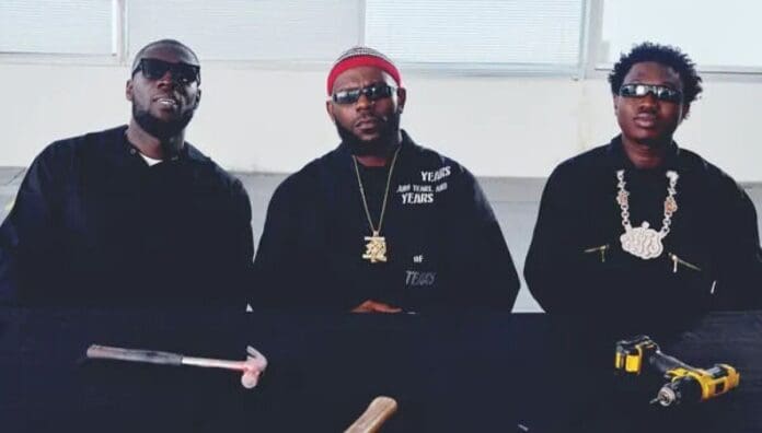 Three men wearing dark clothing and sunglasses sit at a table with a hammer and a yellow power drill in front of them, discussing top Afrobeats songs set to dominate August 2025.