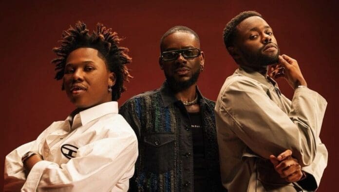 Three men stand together against a brown backdrop, ready to stream the latest Afrobeats songs. The man on the left wears a white shirt, the middle one sports sunglasses and a dark jacket, and the rightmost has on a light coat—September 2025 vibes.