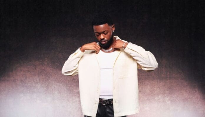 A man stands against a textured background, wearing a light-colored shirt over a white tank top, adjusting his collar and looking down—capturing the quiet mood of Lojay’s XOXO and emotional preservation.