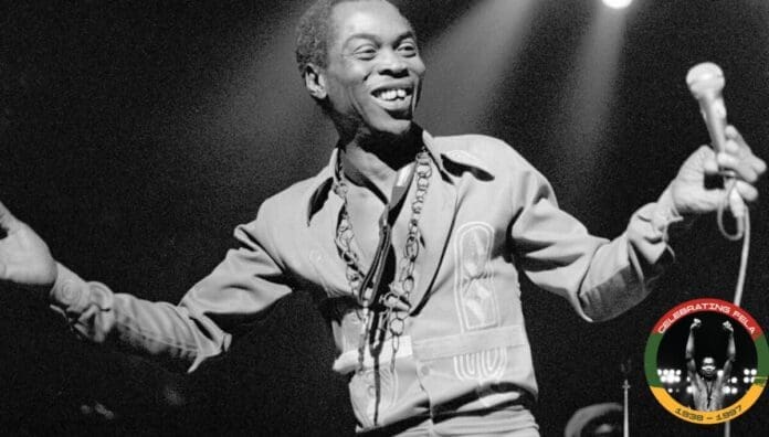 A man smiling and holding a microphone on stage, wearing a patterned shirt inspired by Fela Kuti; there is a circular logo in the bottom right corner, hinting at an Afrobeat vibe.