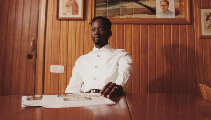 A man in a white shirt sits at a wooden table with a newspaper in front of him, against a wood-paneled wall with framed pictures, evoking the relaxed vibe of Mr Eazi enjoying Afrobeats at Maison Rouge.