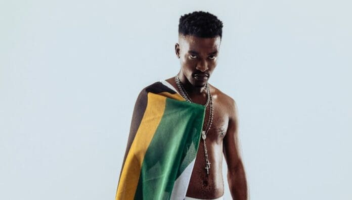A man with short hair and necklaces stands shirtless, draped in a green, gold, and black flag against a plain light background—capturing the vibrant spirit featured in OkayAfrica’s Top African Songs.