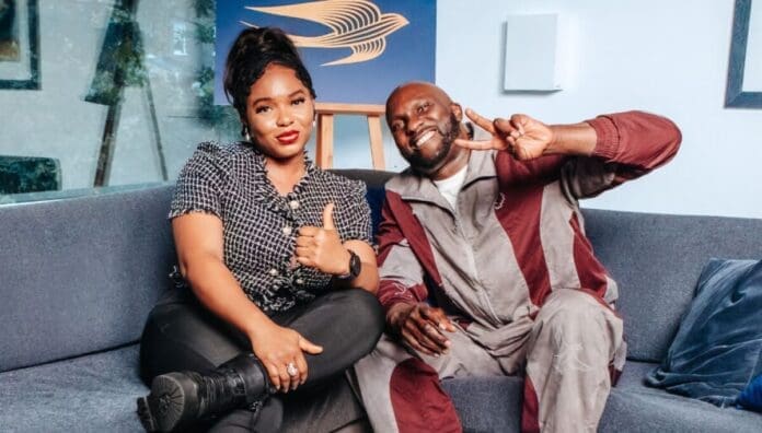 Two people sit on a couch; the woman gives a thumbs up and the man makes a peace sign with his fingers. Both are smiling at the camera, sharing Swift Conversations about Afrobeats.