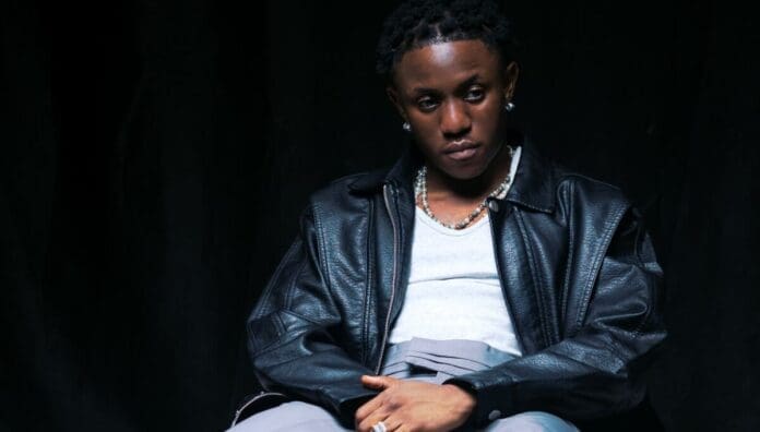 A person wearing a black leather jacket, gray pants, and a white shirt sits against a dark background, looking slightly downward with a neutral expression—evoking the cool confidence often seen on Top African songs playlists by OkayAfrica.