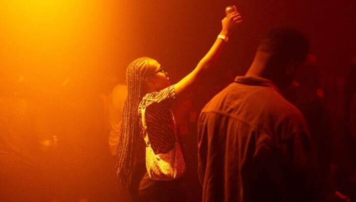 A person with braids raises a drink in a dimly lit, orange-hued crowded room, as Amapiano beats pulse through the space, surrounded by other people.