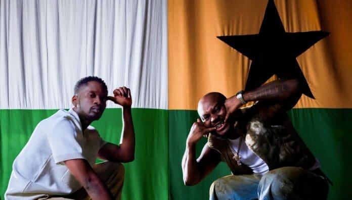 Two men pose in front of a large flag with green, white, and orange stripes and a black star on the orange stripe, capturing the vibrant spirit found in African music featured on OkayAfrica.