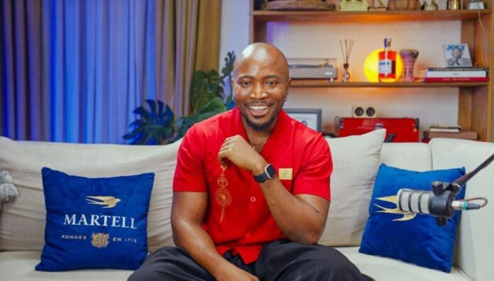 A man in a red shirt sits on a beige sofa with blue Martell pillows, smiling at the camera. Shelves with books, bottles, and decor are in the background—a cozy setting for an Afrobeats Intelligence Season 5 interview.