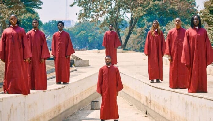 Eight people in matching red choir robes stand spaced apart outdoors on a concrete walkway, performing choir renditions with an Afrobeats twist. Trees and buildings are visible in the background, creating a lively scene reminiscent of OkayAfrica features.