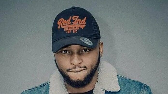 A man wearing a black "Red Ind" cap, a blue denim jacket with a light-colored shearling collar, and a short beard stands against a plain background, channeling cool Amapiano vibes like Sir Trill to impress his Spotify followers.