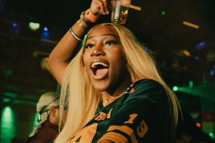 A woman with long blonde hair cheers with unfiltered joy, drink raised above her head, wearing a dark sports jersey in a lively, dimly lit setting that captures the vibrant energy of club culture.