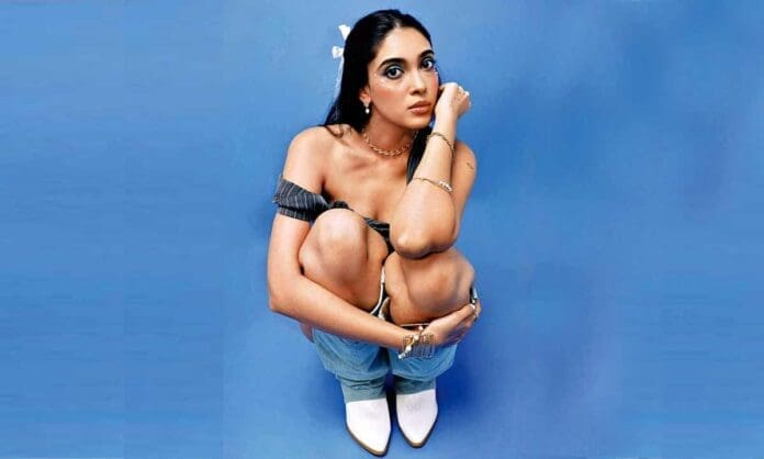 A woman with long hair sits on the floor against a blue background, hugging her knees in a Lonely pose, wearing an off-shoulder top, jeans, and white boots—capturing a mood reminiscent of an afro-pop album cover by Khevna.
