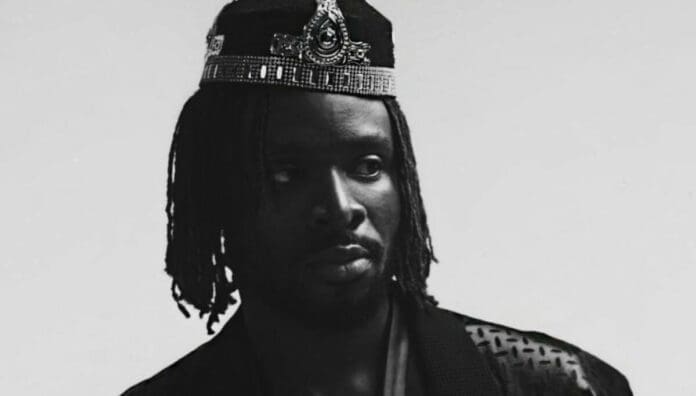 A person with medium-length dreadlocks wears a decorative crown and patterned clothing, looking slightly to the side against a plain background—evoking OkayAfrica style and the vibrant flair of African names.