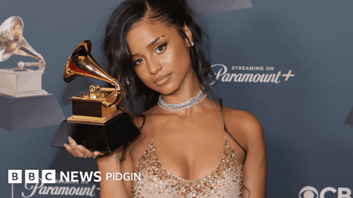 A woman in a gold dress holds a Grammy award and poses for a photo at an event, celebrating her spot among the 2026 Grammy Awards winners, with a blue backdrop featuring logos behind her.