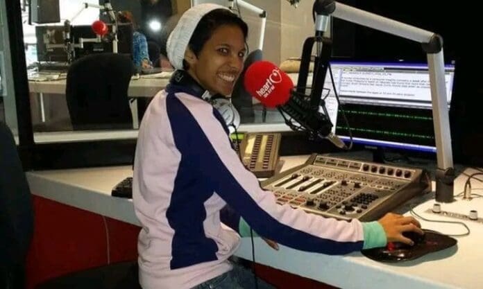 A person wearing a headset and light jacket sits at a radio station desk, speaking into a microphone and using a computer to explore South African music on the Plugm8 music submission platform.