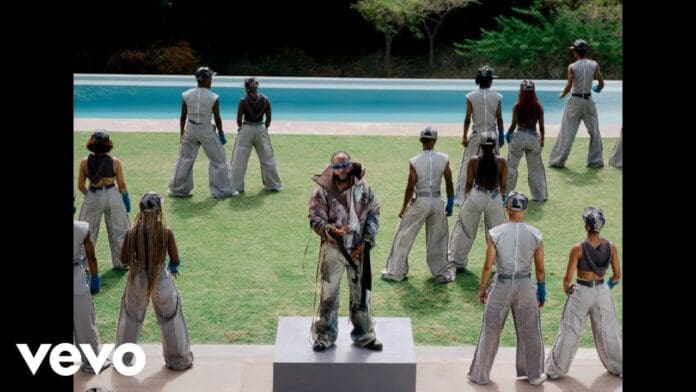 A performer stands on a platform in front of a group of dancers wearing gray outfits and gloves, with a pool and trees in the background, capturing the energy of Davido’s Official Video for KANTE.