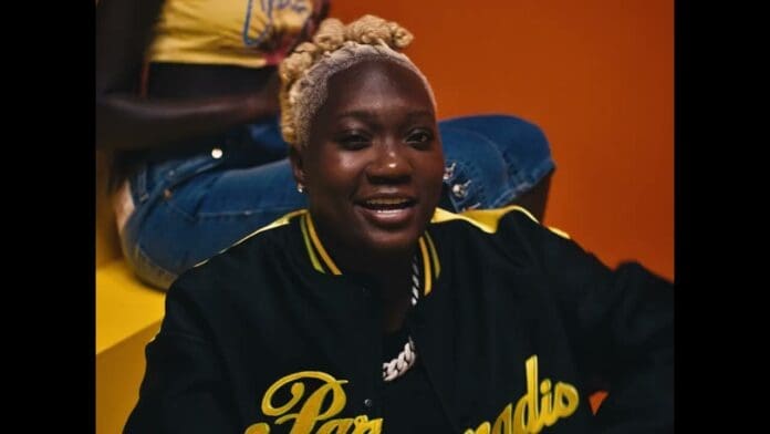 A person with blonde braided hair, wearing a black jacket with yellow lettering, sits in front of an orange background—channeling vibes from Darkoo's "Like Dat" Official Video. Another person in denim shorts is partially visible behind them.