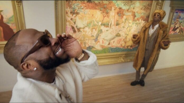 Two men wearing fur coats and sunglasses pose in an art gallery with ornate framed paintings on the walls, giving off serious MAVO vibes.
