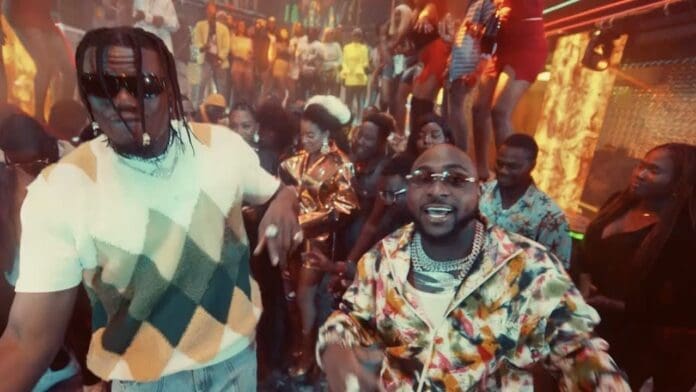 Two men stand in front of a lively crowd at a nightclub, with colorful lights and people dancing in the background, as Davido and Pheelz electrify the atmosphere with their dynamic presence.