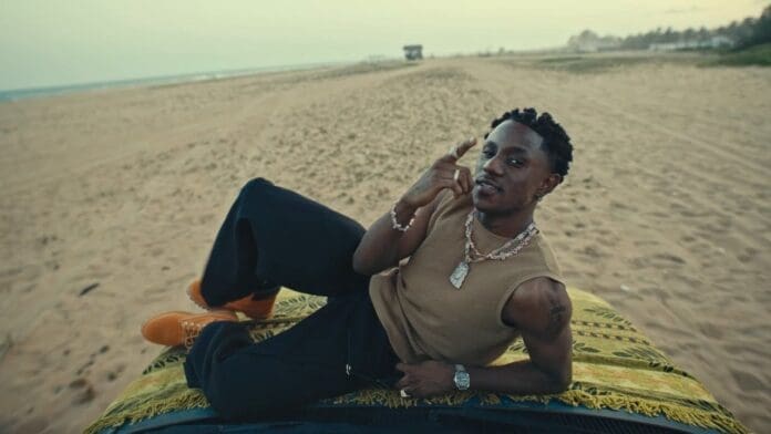 A man lies on his side atop a patterned blanket on a car, on a sandy beach, gesturing with one hand—channeling the laid-back vibes of Victony.