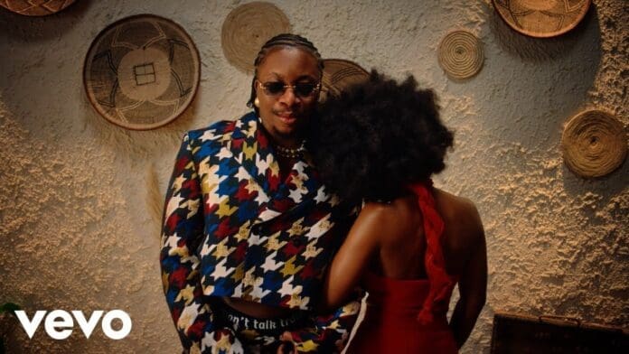 A person in a colorful suit and sunglasses stands next to another in a red dress, leaning on their shoulder, with woven baskets behind them. The Vevo logo is visible—scene from Oxlade’s OMALICHA Official Video.