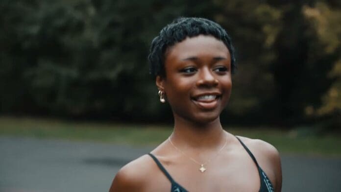 1770159917_maxresdefault.jpg A person with short hair and hoop earrings smiles outdoors, wearing a necklace and a sleeveless top. Trees and greenery are blurred in the background, giving an Official Music Video vibe similar to Mightyyout or Davido’s style.