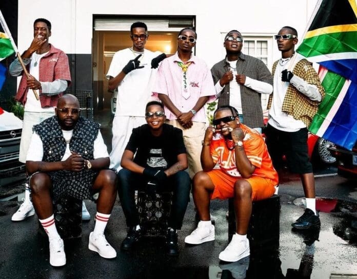 Eight people pose together outdoors in front of a building, with two holding South African flags; several wear sunglasses and streetwear, celebrating their love for Amapiano.